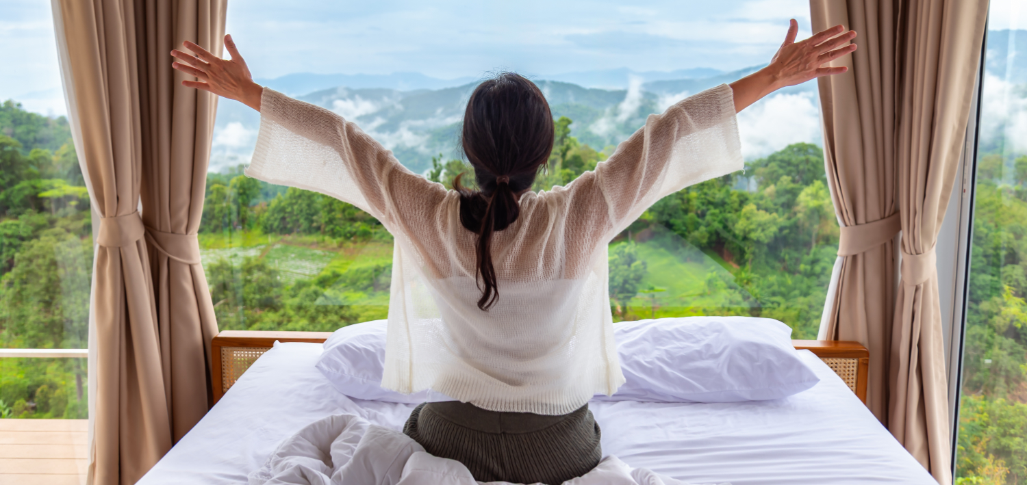 Happy asian woman wakes up  raised open arms on bed with panoramic morning nature mountain and fog view background.
