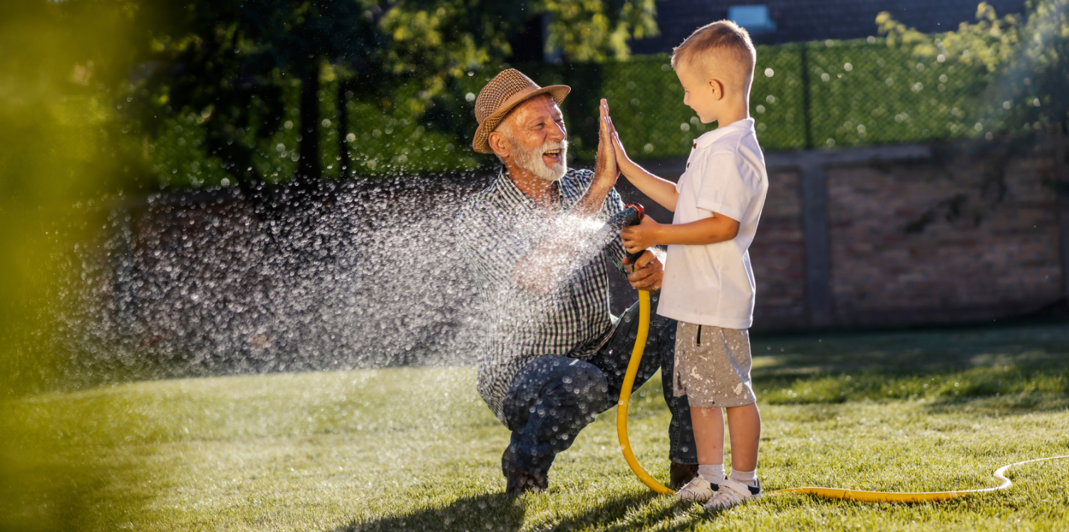 Happy senior man giving high five for team work to little boy while watering lawn in backyard.